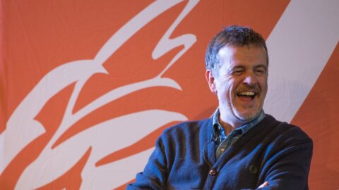A man laughing on stage, with the Stratford Literary Festival logo in the background.