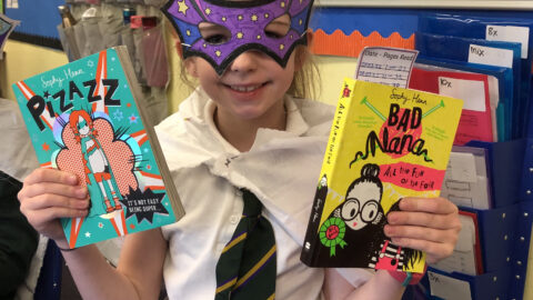 Child wearing a mask and holding up two books in a school classroom.